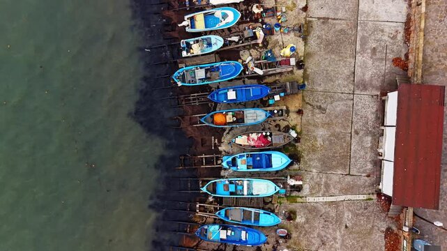 Aerial view of fishing boats on wooden slipway. Overhead drone shot of blue wooden boats on rails beside a concrete pier and red roofed building.