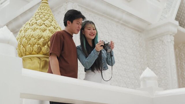 Happy young Asian couple taking selfie video and exploring beautiful white temple architecture. Cheerful romantic tourist lovers enjoying cultural sightseeing summer vacation.