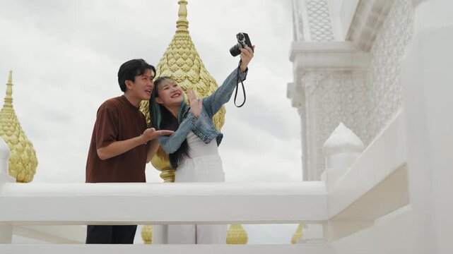 Happy young Asian couple taking selfie video and exploring beautiful white temple architecture. Cheerful romantic tourist lovers enjoying cultural sightseeing summer vacation.