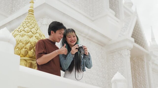 Happy young Asian couple taking selfie video and exploring beautiful white temple architecture. Cheerful romantic tourist lovers enjoying cultural sightseeing summer vacation.