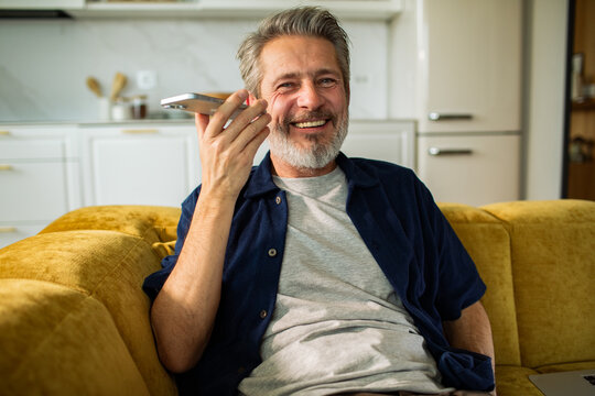 Smiling mature man using smartphone voice assistant on sofa at home