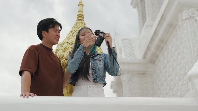 Happy young Asian couple taking selfie video and exploring beautiful white temple architecture. Cheerful romantic tourist lovers enjoying cultural sightseeing summer vacation.