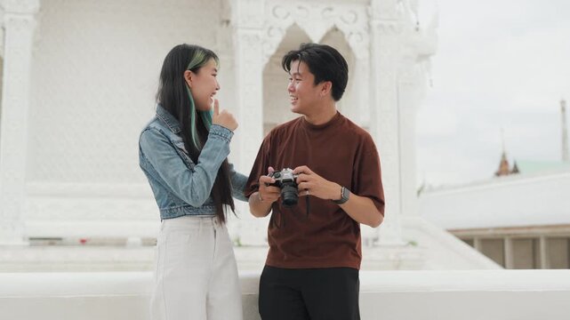 Attractive young Asian couple looking at digital camera pictures at beautiful white temple. Happy romantic tourist travelers enjoying historic sightseeing summer holiday vacation.