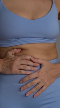 Young woman touching bloated belly in blue sportswear, showing abdominal fat, swelling and stomach discomfort, digestion and body problem concept on gray background