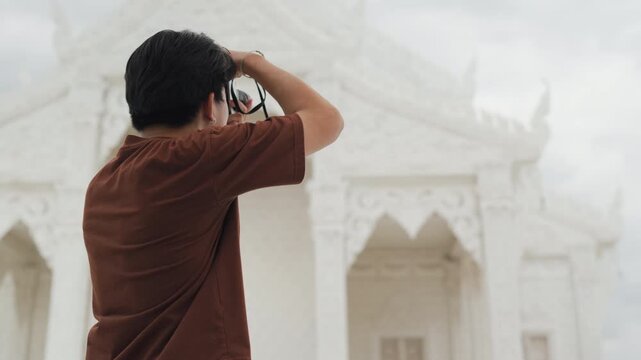 Handsome young Asian male tourist taking beautiful photos with digital camera at white temple. Cheerful solo traveler enjoying cultural sightseeing adventure on summer vacation.