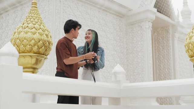Attractive young Asian couple looking at digital camera pictures at beautiful white temple. Happy romantic tourist travelers enjoying historic sightseeing summer holiday vacation.