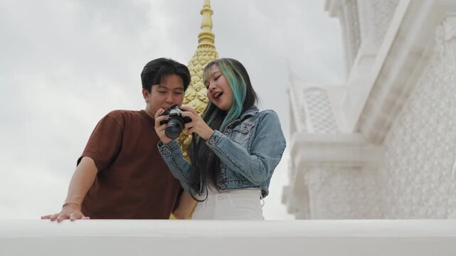 Attractive young Asian couple looking at digital camera pictures at beautiful white temple. Happy romantic tourist travelers enjoying historic sightseeing summer holiday vacation.