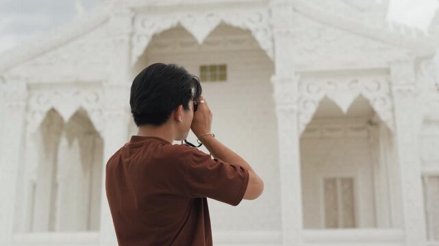 Handsome young Asian male tourist taking beautiful photos with digital camera at white temple. Cheerful solo traveler enjoying cultural sightseeing adventure on summer vacation.