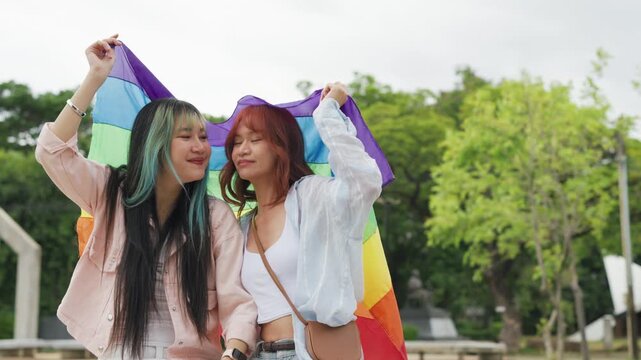 Beautiful Asian lesbian couple holding a rainbow flag and embracing in green park. Cheerful young women celebrating LGBTQ pride month equality and expressing romantic love.