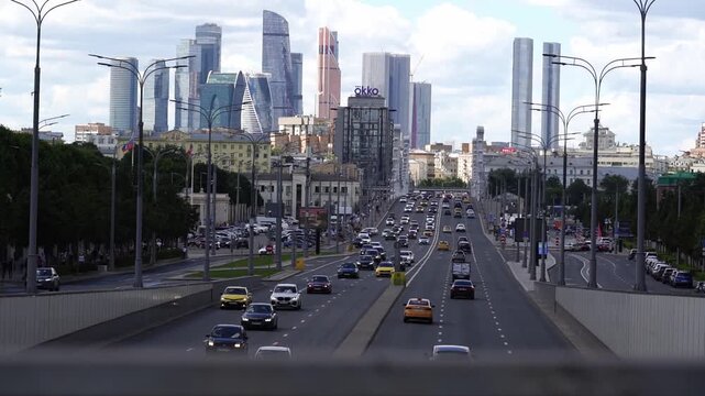 City streets bustle with cars and distant skyscrapers. Vehicles move steadily along wide roads lined with trees. Urban skyline dominates the background under a cloudy sky. People drive focused