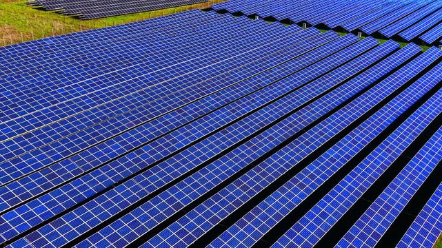 Panoramic aerial view of solar farm rows. Diagonal panel arrays extend across a hillside toward another array in the distance.
