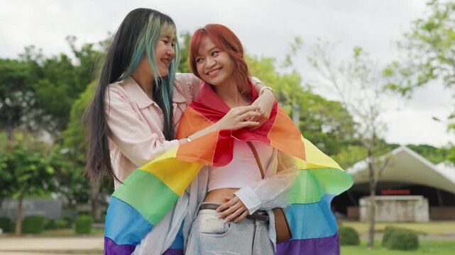 Beautiful Asian lesbian couple holding a rainbow flag and embracing in green park. Cheerful young women celebrating LGBTQ pride month equality and expressing romantic love.