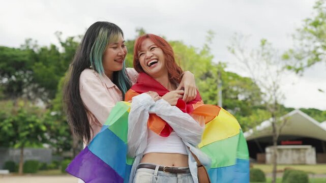 Beautiful Asian lesbian couple holding a rainbow flag and embracing in green park. Cheerful young women celebrating LGBTQ pride month equality and expressing romantic love.