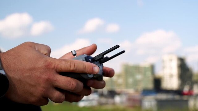 Hands grip remote control firmly outdoors. Sky and buildings blur softly behind. Focus on device and steady grip. Controlled flight operation underway. Ideal for drone or tech hobbyist scenes
