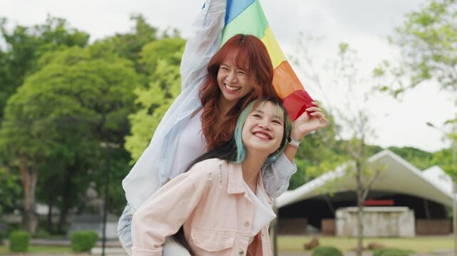 Beautiful Asian lesbian couple holding a rainbow flag and embracing in green park. Cheerful young women celebrating LGBTQ pride month equality and expressing romantic love.