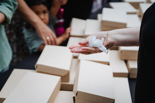 Closeup volunteer hands out crumpled tissue beside stacked cardboard boxes at donation table, with blurred recipients reaching in background and soft natural light creating calm humanitarian mood