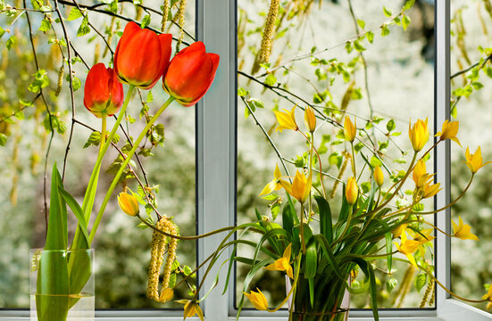 Flowers are arranged in vases by a window with greenery in the background on a sunny day