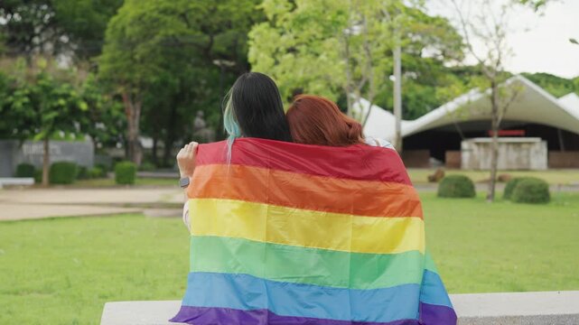 Beautiful Asian lesbian couple holding a rainbow flag and embracing in green park. Cheerful young women celebrating LGBTQ pride month equality and expressing romantic love.