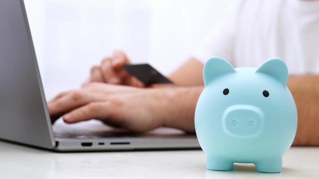 Blue piggy bank sits on desk. Hands type on laptop, hold card. Focus is on saving, spending, digital finance. Perfect for budgeting, online shopping, finance themes. No animals, no plants