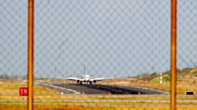 Airplane lands behind rusty chain-link fence. Runway stretches under clear, sunlit sky. Vehicles visible in distance, motion blurred. Ideal for travel, aviation, or security concept visuals