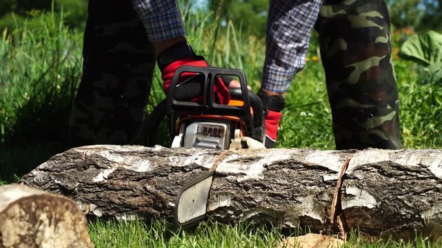 Person cuts log with chainsaw on green grass. Red gloves grip machine firmly. Camo pants and plaid shirt cover sturdy build. Chainsaw blade spins, slicing wood