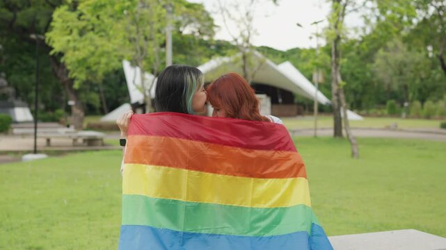 Beautiful Asian lesbian couple holding a rainbow flag and embracing in green park. Cheerful young women celebrating LGBTQ pride month equality and expressing romantic love.