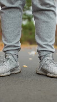 Gray fabric pants and sneakers visible as feet occupy. Paved surface hosts fallen leaves as feet remain still. Stationary figure wears light gray knit footwear surrounded