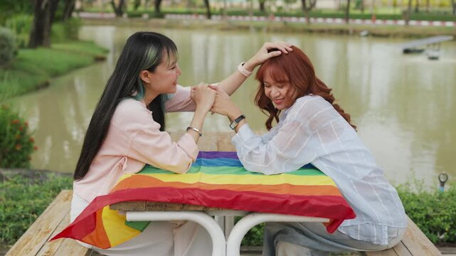 Beautiful Asian lesbian couple holding a rainbow flag and embracing in green park. Cheerful young women celebrating LGBTQ pride month equality and expressing romantic love.