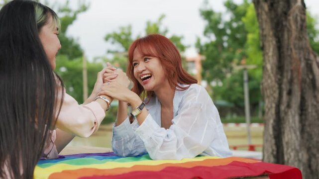 Happy young Asian lesbian couple sitting on a rainbow flag enjoying romantic summer picnic in park. Beautiful LGBTQ women sharing lovely weekend date and relaxing lifestyle.