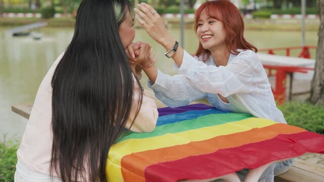 Happy young Asian lesbian couple sitting on a rainbow flag enjoying romantic summer picnic in park. Beautiful LGBTQ women sharing lovely weekend date and relaxing lifestyle.