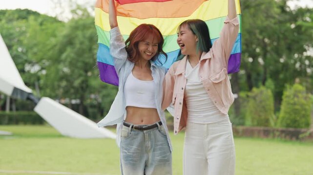 Happy young Asian lesbian couple sitting on a rainbow flag enjoying romantic summer picnic in park. Beautiful LGBTQ women sharing lovely weekend date and relaxing lifestyle.