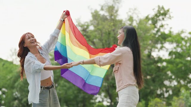 Happy young Asian lesbian couple sitting on a rainbow flag enjoying romantic summer picnic in park. Beautiful LGBTQ women sharing lovely weekend date and relaxing lifestyle.