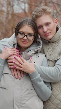 Young couple holds hands while standing side by side. Quilted jacket is worn by male subject in outdoor scene. Outdoor setting shows blurred trees and natural environment