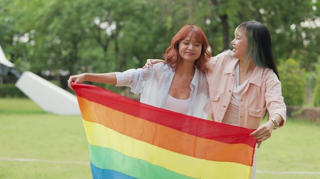 Happy young Asian lesbian couple sitting on a rainbow flag enjoying romantic summer picnic in park. Beautiful LGBTQ women sharing lovely weekend date and relaxing lifestyle.