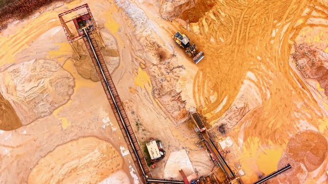 Industrial conveyor tower at sand quarry. Overhead composition highlights a red metal conveyor structure beside an idle wheel loader.
