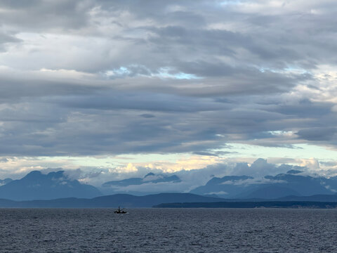 coastal mountains of southwestern Canada at dusk with sunset colors on distant clouds over land and a layer of gray clouds over the ocean, with a fishing boat looking very small in the vast landscape