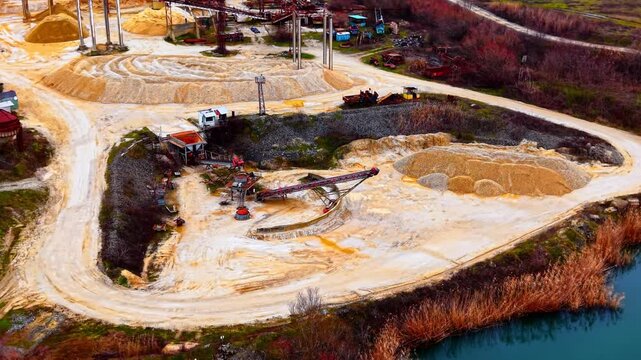 Aerial view of gravel processing site by lake, Bulgaria. Wide angle shows conveyors, sand mounds, a dry reed bed, and distant water reflections.