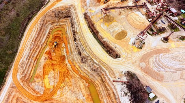 Sand quarry pit with excavator near forest. Aerial view of terraced orange sand banks, a winding stream, and processing equipment.