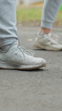 Pair of legs with one foot ahead both steps visible. Shoes vary between new and used condition with gray pants. Outdoor setting with blurred greenery behind paved surface
