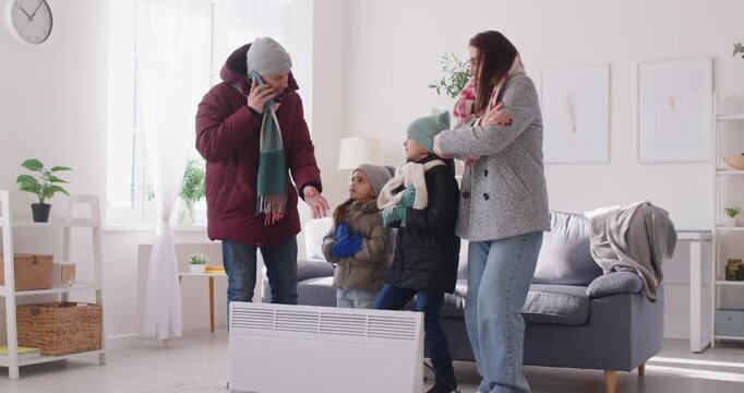 Parents and small children freezing at home wearing winter warm clothes near radiator heater, badly shivering, cold at home, call apartment management company to complain about low temperature