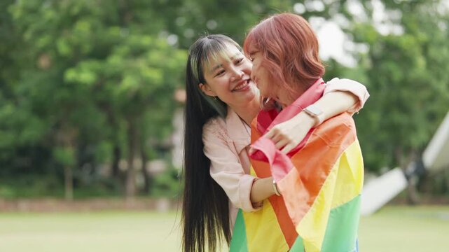 Happy young Asian lesbian couple sitting on a rainbow flag enjoying romantic summer picnic in park. Beautiful LGBTQ women sharing lovely weekend date and relaxing lifestyle.