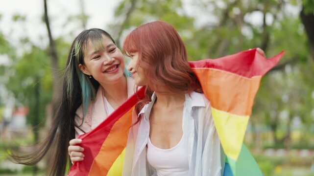 Happy young Asian lesbian couple sitting on a rainbow flag enjoying romantic summer picnic in park. Beautiful LGBTQ women sharing lovely weekend date and relaxing lifestyle.