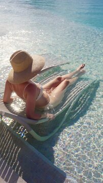 Vertical Screen: Woman wearing straw hat and white bikini lounges on submerged pool chair in Bora Bora. Crystal clear turquoise water sparkles in sunlight as she relaxes at luxury tropical resort.