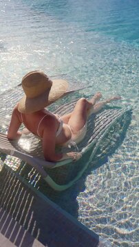 Vertical Screen: Woman in white bikini and wide-brim straw hat relaxes on submerged pool hammock, crystal clear turquoise water sparkling under bright tropical sunlight at luxury resort destination.