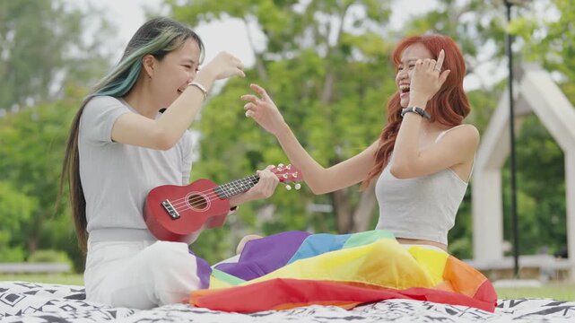 Happy young Asian lesbian couple sitting on a rainbow flag enjoying romantic summer picnic in park. Beautiful LGBTQ women sharing lovely weekend date and relaxing lifestyle.