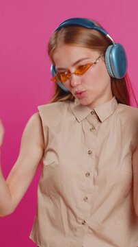 Happy woman in headphones listening music and dancing disco fooling around having fun expressive gesticulating hands relaxing on party. Blonde lady in sunglasses isolated on studio pink background