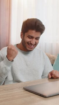 Young Indian man sitting at table with laptop, smiling after completing successful project, clenching fist, closing laptop, and celebrating. Hispanic Hindu guy relaxing on sofa in living room at home.