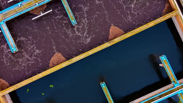 Aerial close up of frothy wastewater treatment pool. Overhead detail of foamy brown aeration basin beside dark clarifier tank with metal walkway.