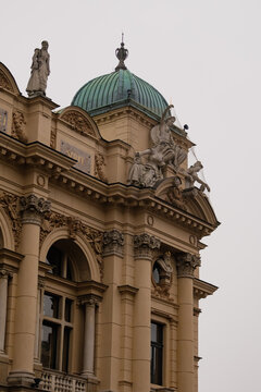 Ornate historic building facade with classical sculptures, columns, and a green copper dome under an overcast sky. Detailed architectural elements and statues highlight European classical architecture