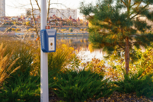 Emergency call box, panic alarm on urban park path beside lake or river, with evergreen plants and golden autumn foliage with city residential landscape background. Public safety and security system
